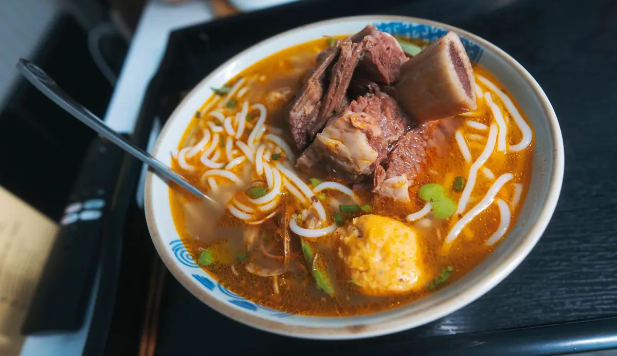 A steaming bowl of local Shacha noodles served in a traditional canteen, featuring rich peanut-based broth and fresh seafood toppings.